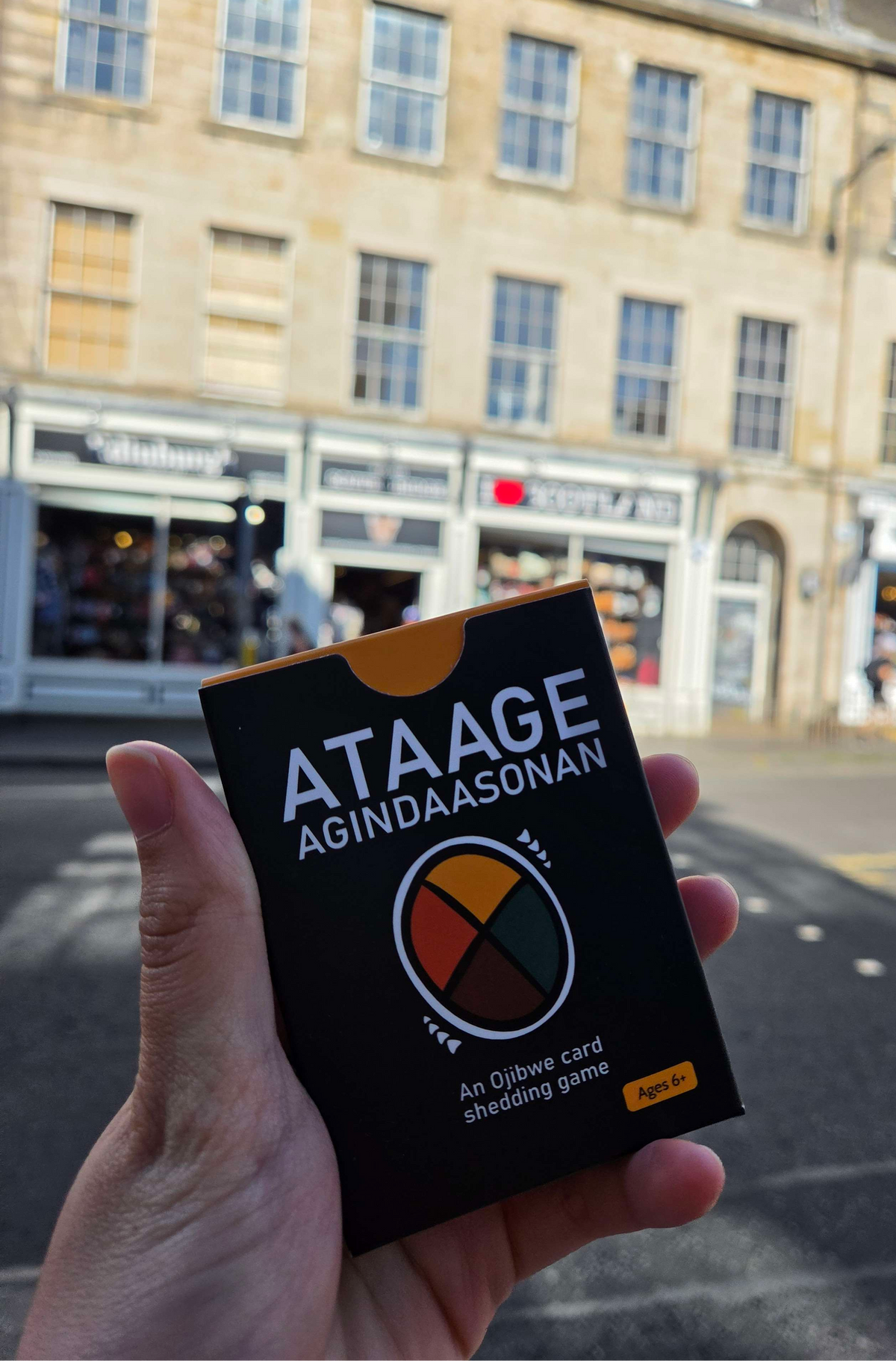 A hand holds a black box of the card game 'ATAAGE AGINDAASONAN' in front of a blurred street scene with buildings and shops.