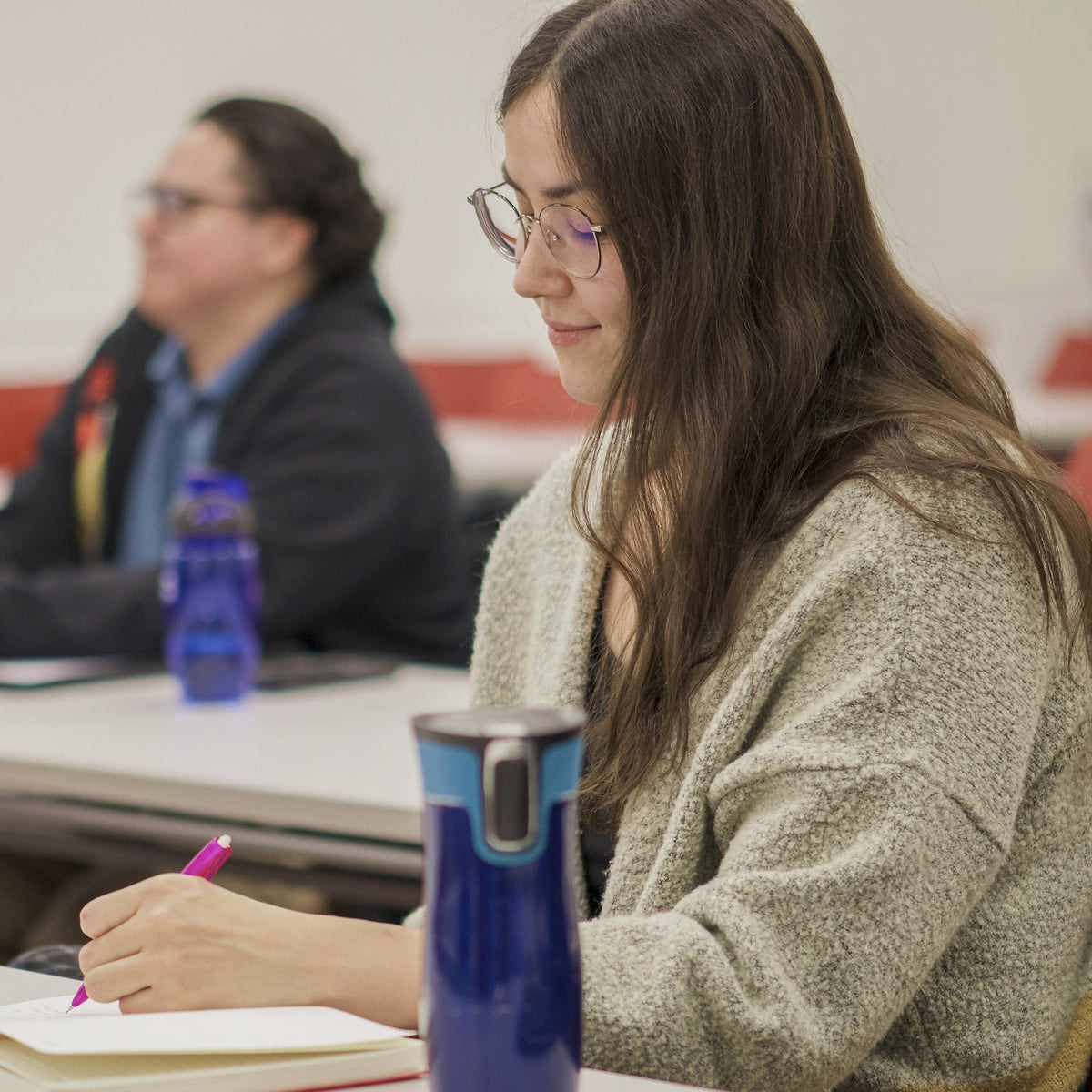 Student sitting at a desk in a classroom, writing in a notebook with a blue water bottle and glasses on the table.
