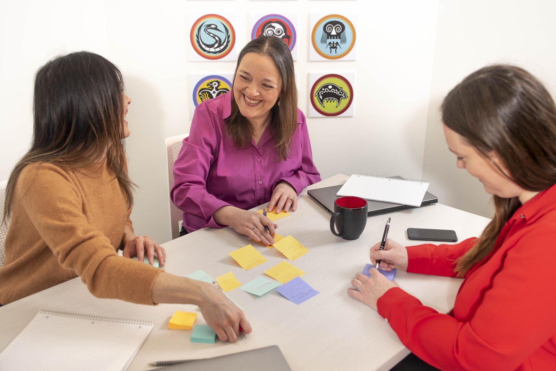 Three women sitting around a table with colorful sticky notes and office supplies.